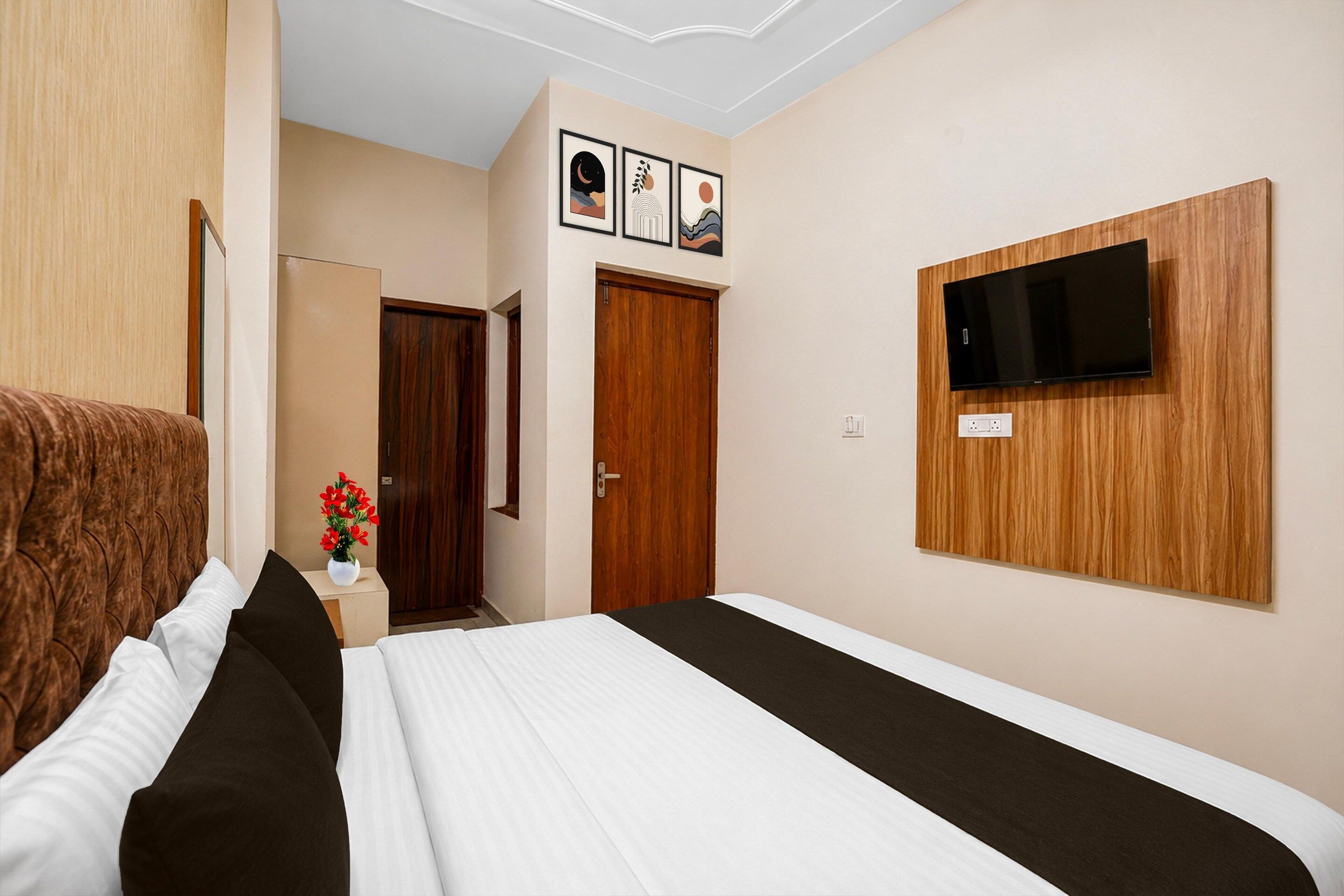 A hotel room with a bed, a TV mounted on the wall, a wooden door, and a small table with a vase of flowers.