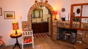 Uma sala de estar com lareira, um banco branco, uma mesa com abajur e uma cozinha visível através de um arco.