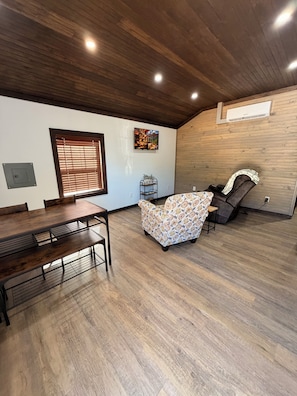 Una habitación amplia con techo de madera, una mesa de comedor, un sillón estampado y una silla de masaje.
