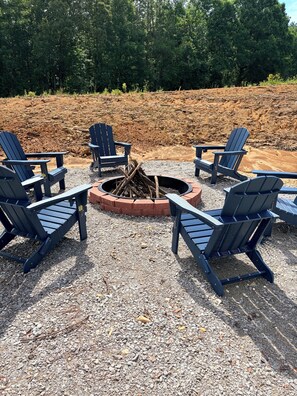 A fire pit surrounded by Adirondack chairs on a gravel surface, with a forested hillside in the background.