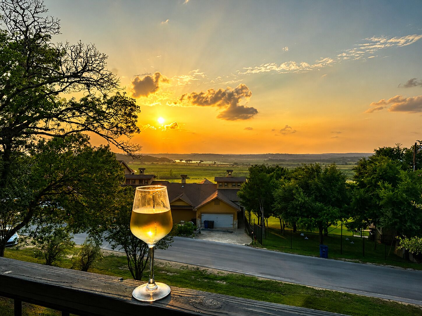 Ein Glas Weißwein auf einer Holzbrüstung mit Blick auf ein Wohngebiet, Häuser und eine Straße bei Sonnenuntergang.