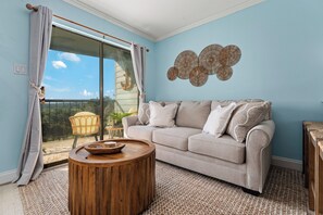Ein Wohnzimmer mit einer Couch, einem runden Holztisch und Blick durch eine Glasschiebetür auf einen Außenbereich mit einem Sessel und Pflanzen.
