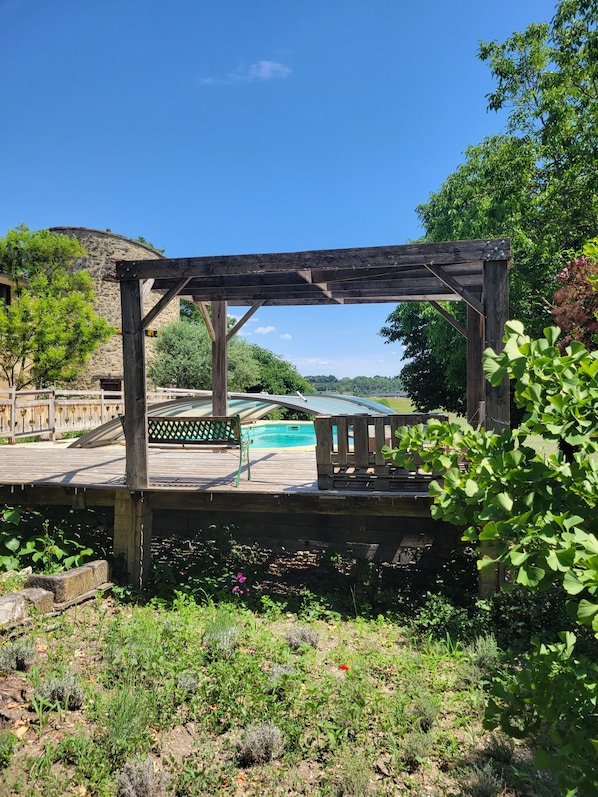 A wooden deck with a pool, surrounded by greenery and a stone wall.