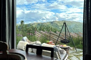 A room with a view of a mountainous landscape, a wooden fence, a hanging light fixture, and a table with a chair.