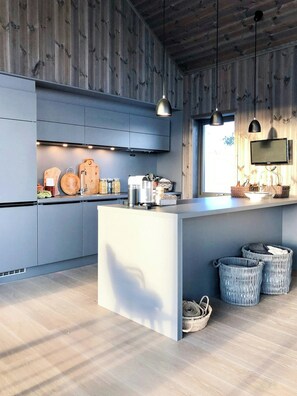 A modern kitchen with grey cabinets, a central island, hanging pendant lights, a TV mounted on the wall, and wicker baskets on the floor.