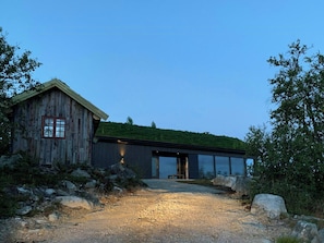 Un bâtiment rustique en bois, agrémenté d’une extension moderne en verre, et entouré d’un paysage naturel.