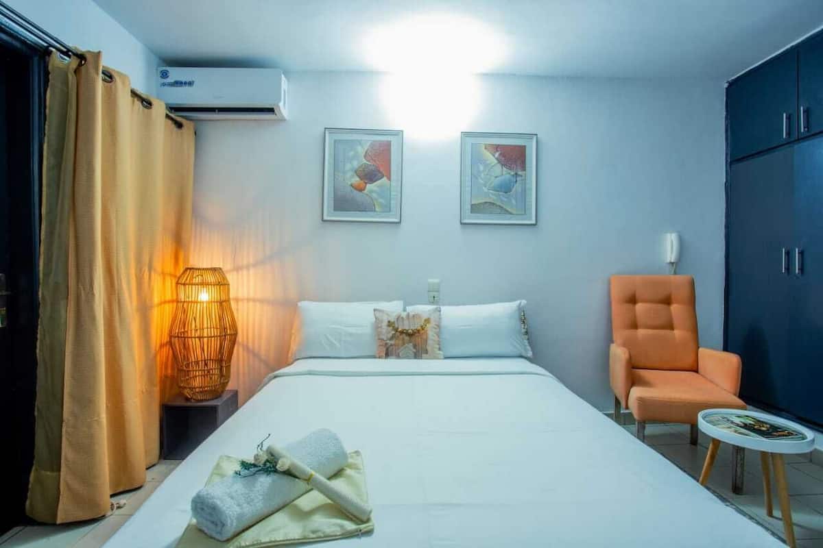 Hotellihuone, jossa on sänky, tuoli, pöytä, lamppu ja kaksi kehystettyä kuvaa seinällä.
