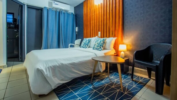 Une chambre d’hôtel avec un lit, une petite table ronde, une chaise, une lampe et un tapis bleu à motifs géométriques.