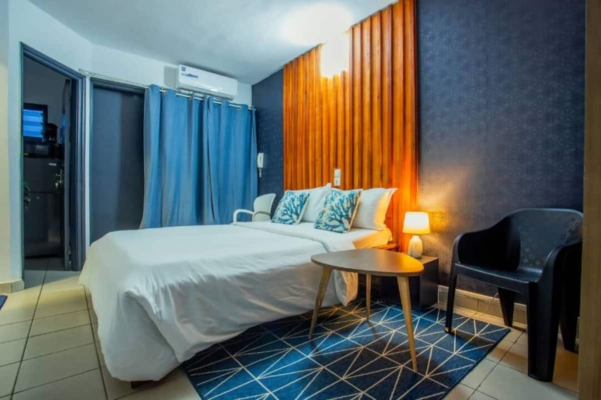 Hotellihuone, jossa on sänky, pieni pyöreä pöytä, tuoli, lamppu ja sininen, geometrisesti kuvioitu matto.