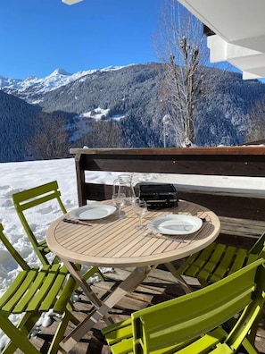 Ein Holztisch mit Stühlen, ein Grill und Blick auf schneebedeckte Berge.
