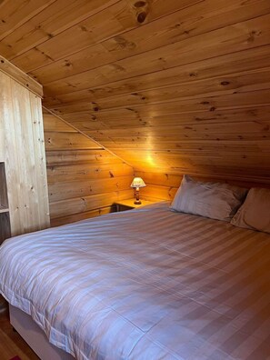 Een slaapkamer in een houten hutje, met een bed, kussens, een nachtkastje en een lamp.
