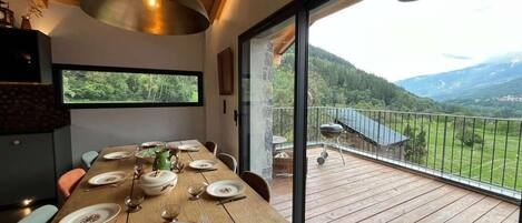 Un comedor con una mesa de madera preparada para cuatro personas, un ventanal con vistas a un paisaje boscoso y un balcón con una parrilla.