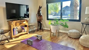Sala de estar com televisão de tela plana, móvel para aparelhos de som de madeira, tapete de yoga, planta em vaso e uma grande janela com vista para o exterior.