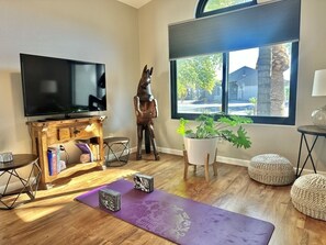 Ein Wohnzimmer mit Flachbildfernseher, einer Holzunterhaltungsecke, einer Yogamatte, einem Topf mit Pflanze und einem großen Fenster mit Blick ins Freie.