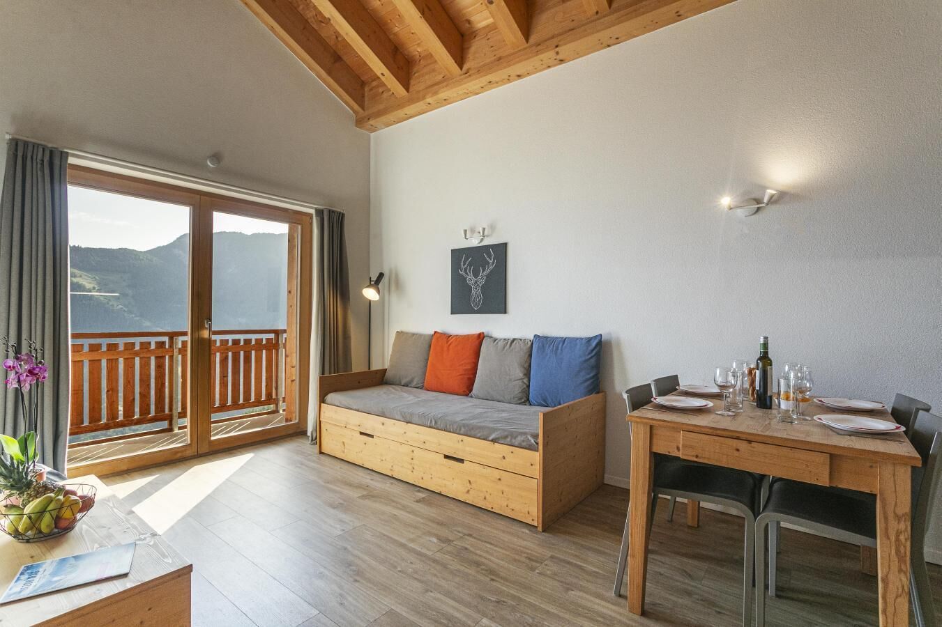 Une pièce avec un plancher en bois, un canapé avec des coussins, une table à manger avec des chaises, une fenêtre donnant sur les montagnes et un luminaire fixé au mur.