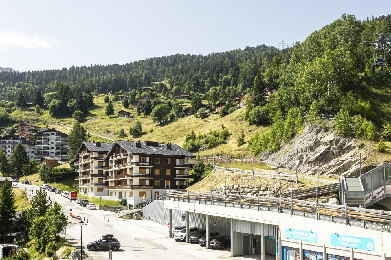 A multi-story hotel with balconies, a parking area, and a ski lift in the background.