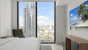Una habitación de hotel moderna con un ventanal grande, una cama, una silla verde, un televisor y un armario de madera.