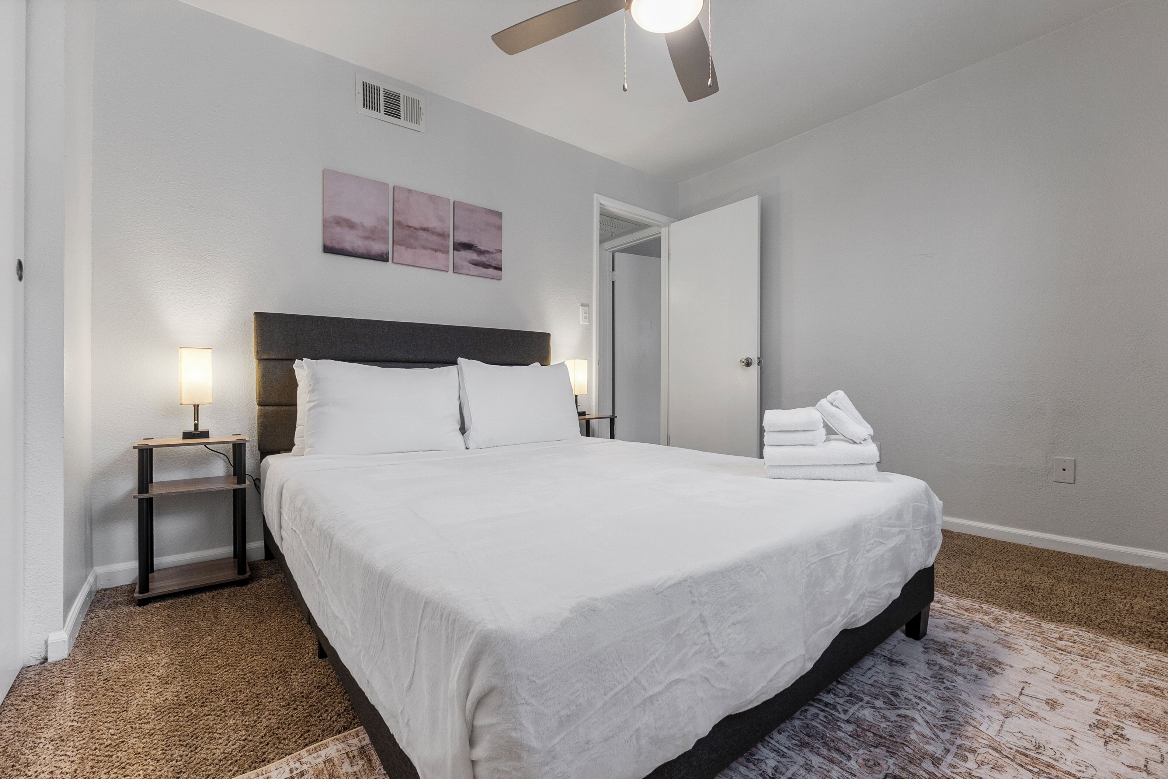 Un dormitorio con cama, mesitas de noche, ventilador de techo y puerta que da a otra habitación.