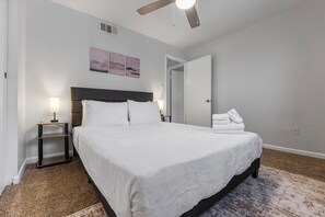 Un dormitorio con cama, mesitas de noche, ventilador de techo y puerta que da a otra habitación.