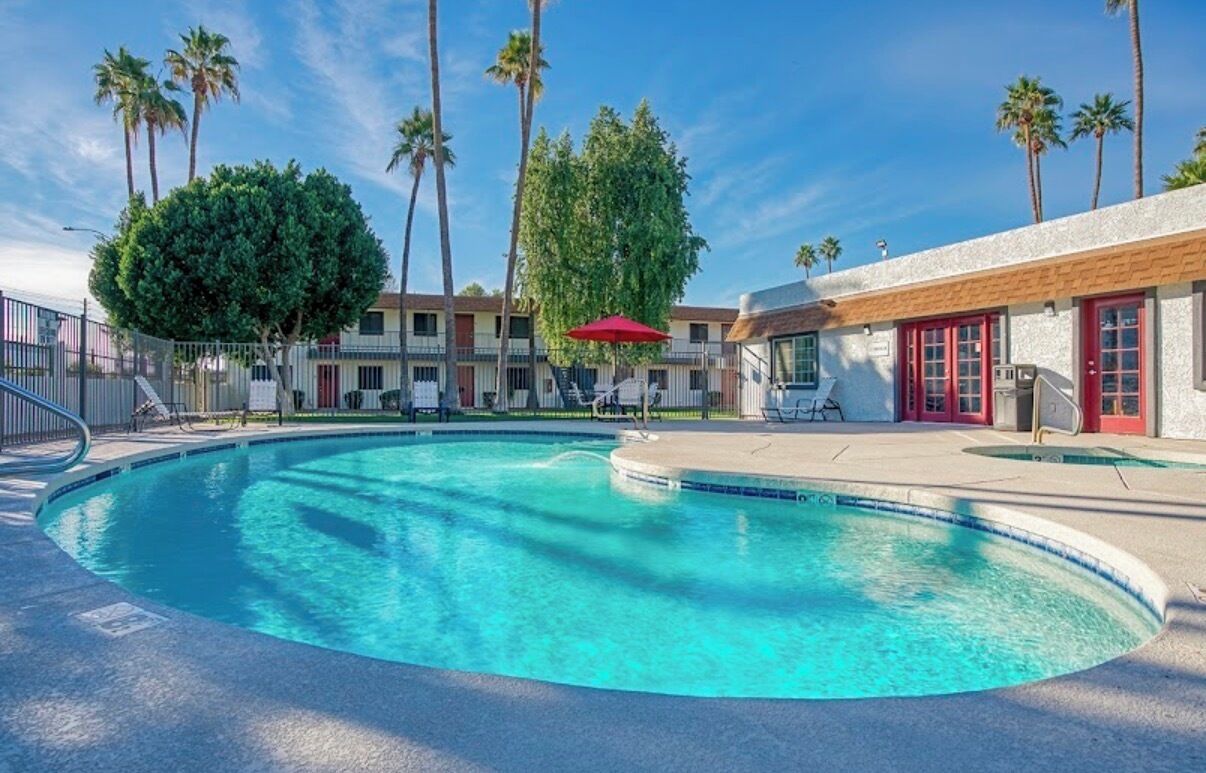 Una piscina con agua azul cristalina, rodeada de palmeras y un edificio con puertas rojas.