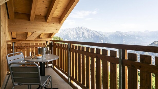 Een houten balkon met een tafel en stoelen, met uitzicht op een bergachtig landschap.