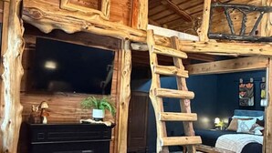 Tunnelmallinen, maalaishenkinen mökin sisustus, jossa on takka, puuseinät, tikapuut ja sänky.