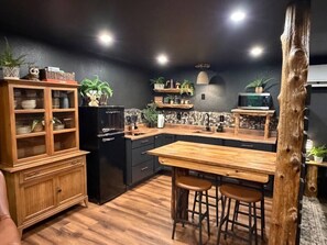 Eine Küche mit Holztisch, Barhocker, Kühlschrank und Regalen mit Pflanzen.