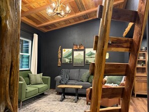 Ein gemütliches Wohnzimmer mit Holzbalkendecke, einem grünen Sofa, einem grauen Sofa, einem Couchtisch und gerahmten Bildern an der Wand.