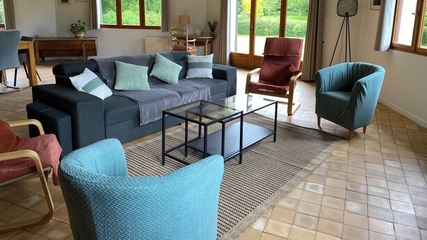 Ein geräumiges Wohnzimmer mit einer großen Couch, einem Couchtisch und zwei Sesseln. Große Fenster mit Vorhängen, ein Essbereich mit Tisch und Stühlen sowie eine Stehlampe sind ebenfalls vorhanden.