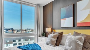 A hotel room with a large window, a bed with a blue blanket, and two abstract paintings on the wall.