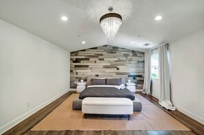 Une chambre à coucher au design moderne, comprenant un lit avec une tête de lit matelassée, un banc au pied du lit, un lustre et un mur texturé derrière le lit.