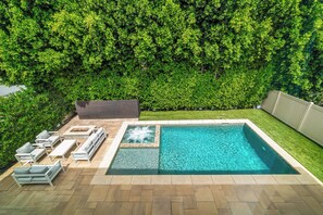 Une piscine rectangulaire avec un jacuzzi, entourée d’une terrasse agrémentée de chaises longues et d’un foyer extérieur.