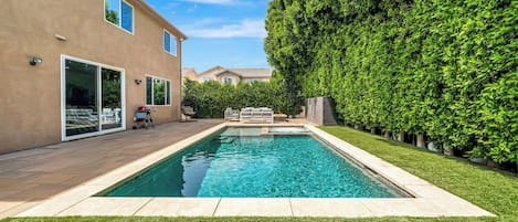 Una piscina con solárium empedrado, rodeada de césped cuidado y un seto alto.