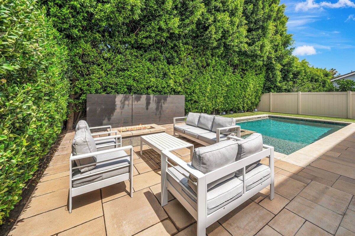 An outdoor patio with a pool, white furniture, and a hedge wall.