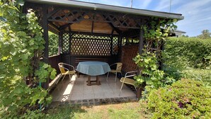 Eine überdachte Terrasse mit Tisch und Stühlen, umgeben von viel Grün.