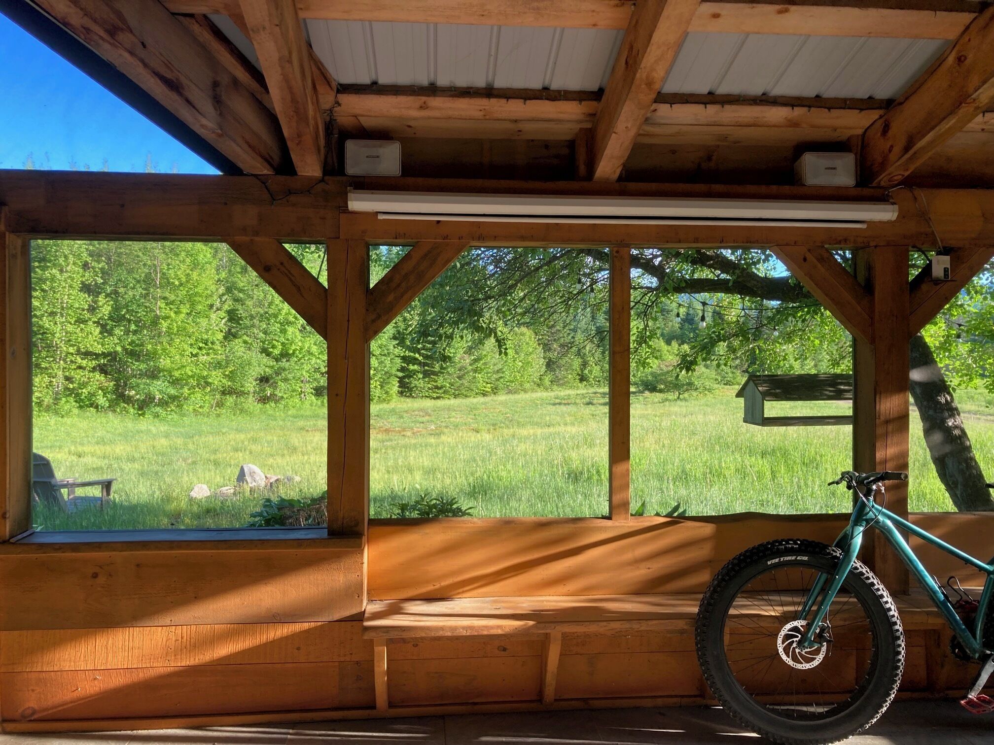 A covered outdoor area with a wooden bench, a bicycle, and a view of a grassy field.