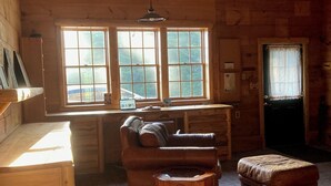 Interior de una cabaña de madera con un sofá, reposapiés y una pequeña mesa. Hay dos ventanas con cortinas, una lámpara en el techo y una puerta con ventanal.