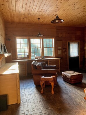 A wooden cabin interior with a sofa, ottoman, and a small table. There are two windows with curtains, a ceiling light, and a door with a window. - Gorgeous 3-bedroom 3 bath Craftsman home north of Mt. Washington (Stark)