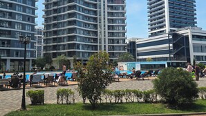 Un entorno urbano moderno con edificios altos, una zona de piscina con tumbonas y un patio pavimentado con mesas y sillas.