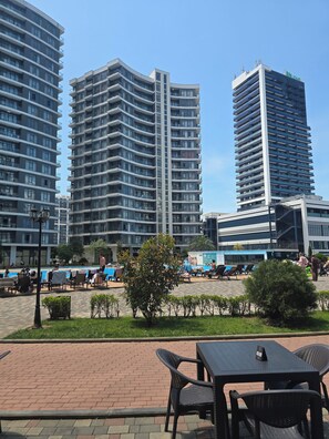 Ein modernes Stadtbild mit Hochhäusern, einem Poolbereich mit Liegestühlen und einer gepflasterten Terrasse mit Tischen und Stühlen.