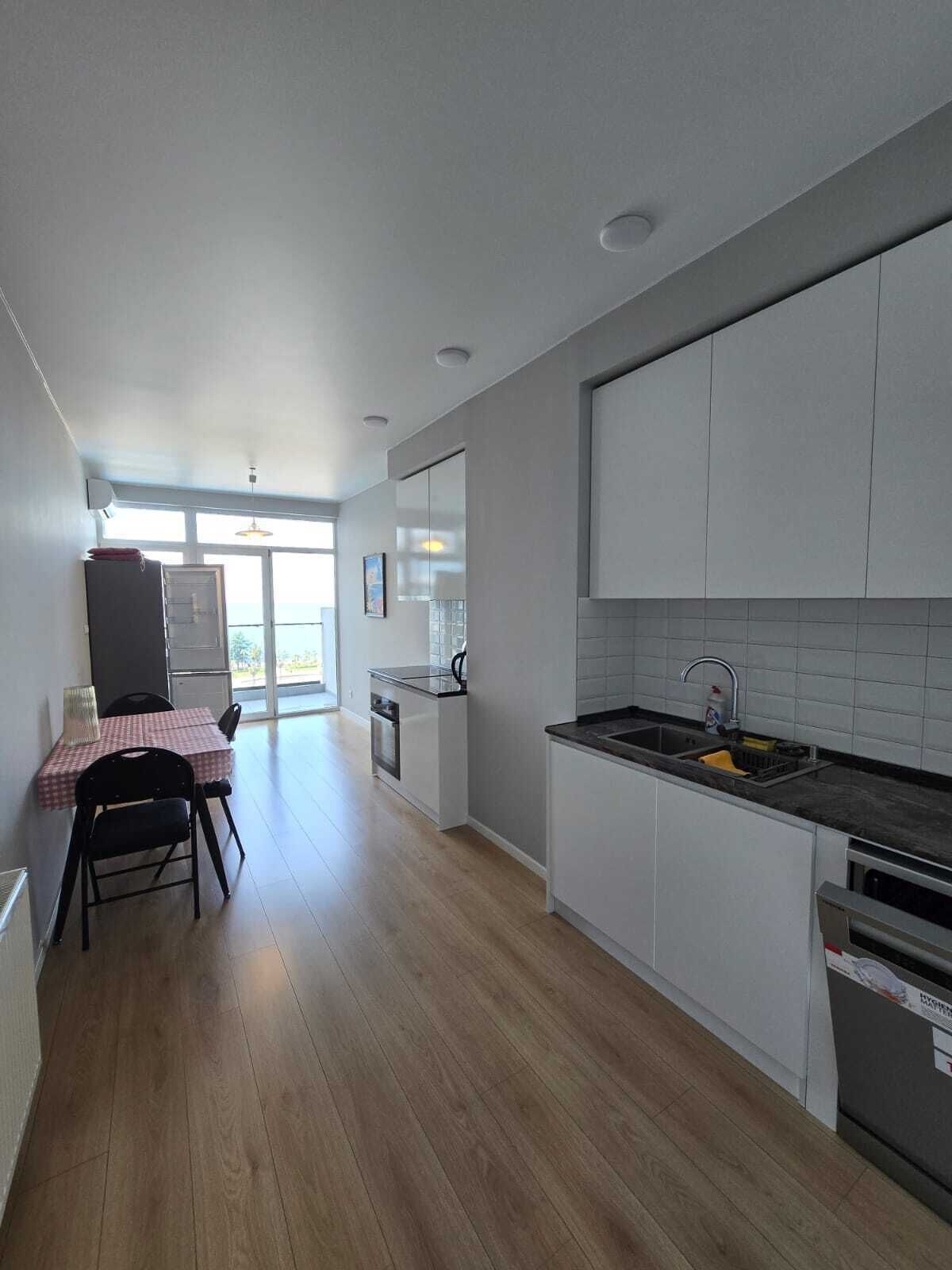 Una cocina moderna con armarios blancos, encimera negra y un lavamanos. Hay un comedor con una mesa y sillas.