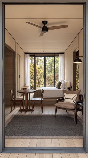 A hotel room with a bed, a desk, two chairs, a ceiling fan, and large windows.