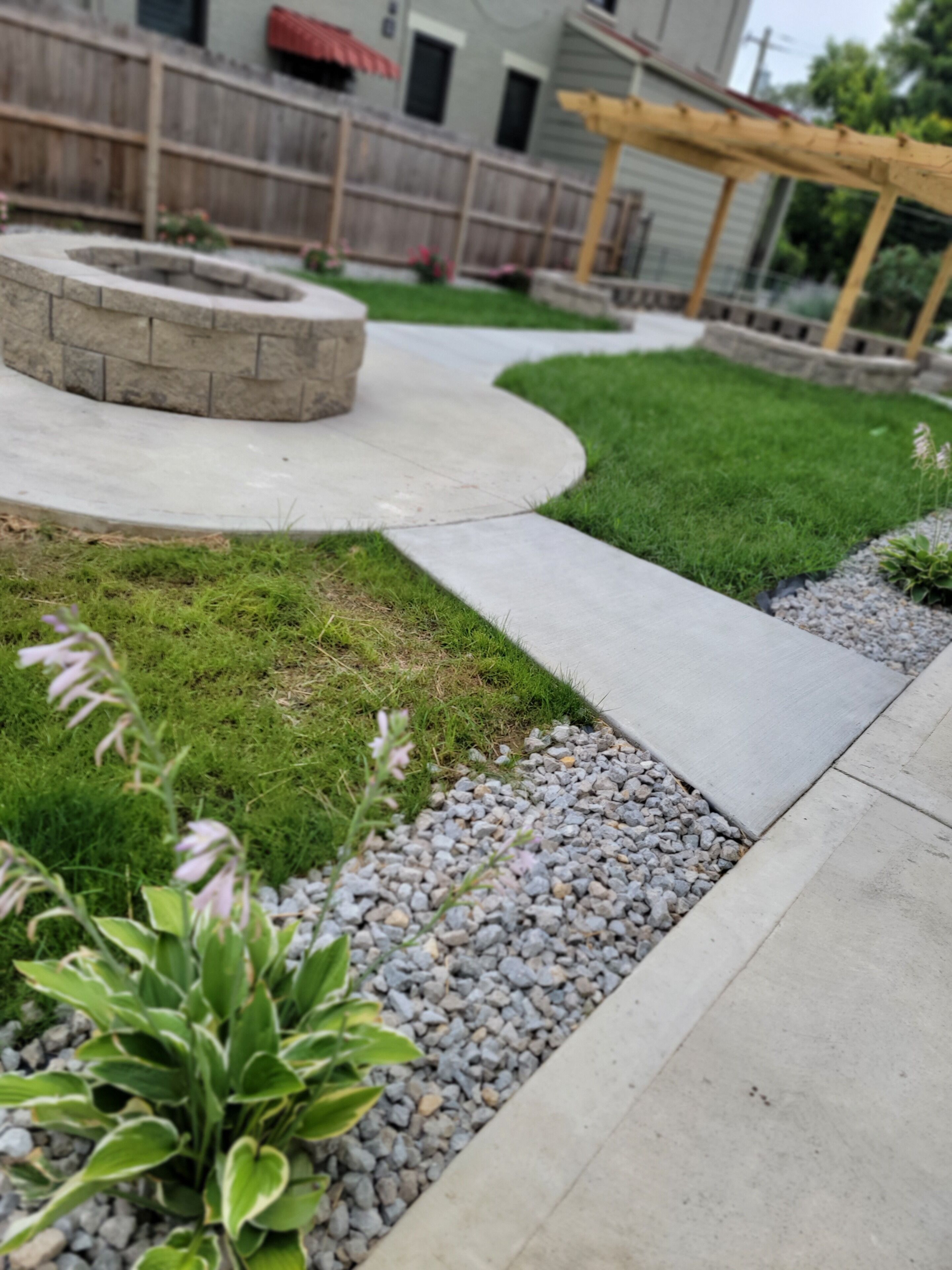 Ein angelegter Garten mit Feuerstelle aus Stein, einem geschwungenen Weg und einer Holzpergola.