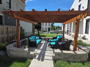 A covered patio with outdoor furniture, a stone border, and a view of a white building in the background. - Cozy 1-bedroom Queen City Urban Escape (Cincinnati)