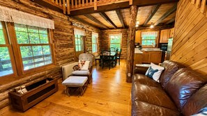 L’intérieur d’une cabane en rondins, avec un plancher en bois, un canapé en cuir, une table à manger avec des chaises et un coin cuisine.