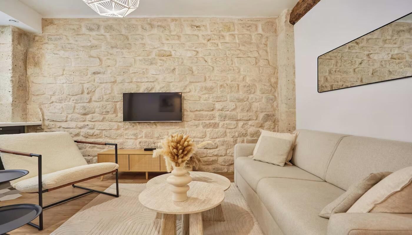 A living room with a stone wall, a flat-screen TV, a sofa, a chair, a small table with a vase, and a wooden side table.