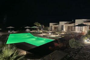 Vista noturna de uma área de piscina com espreguiçadeiras e guarda-sóis, cercada por jardins bem cuidados e edifícios modernos.