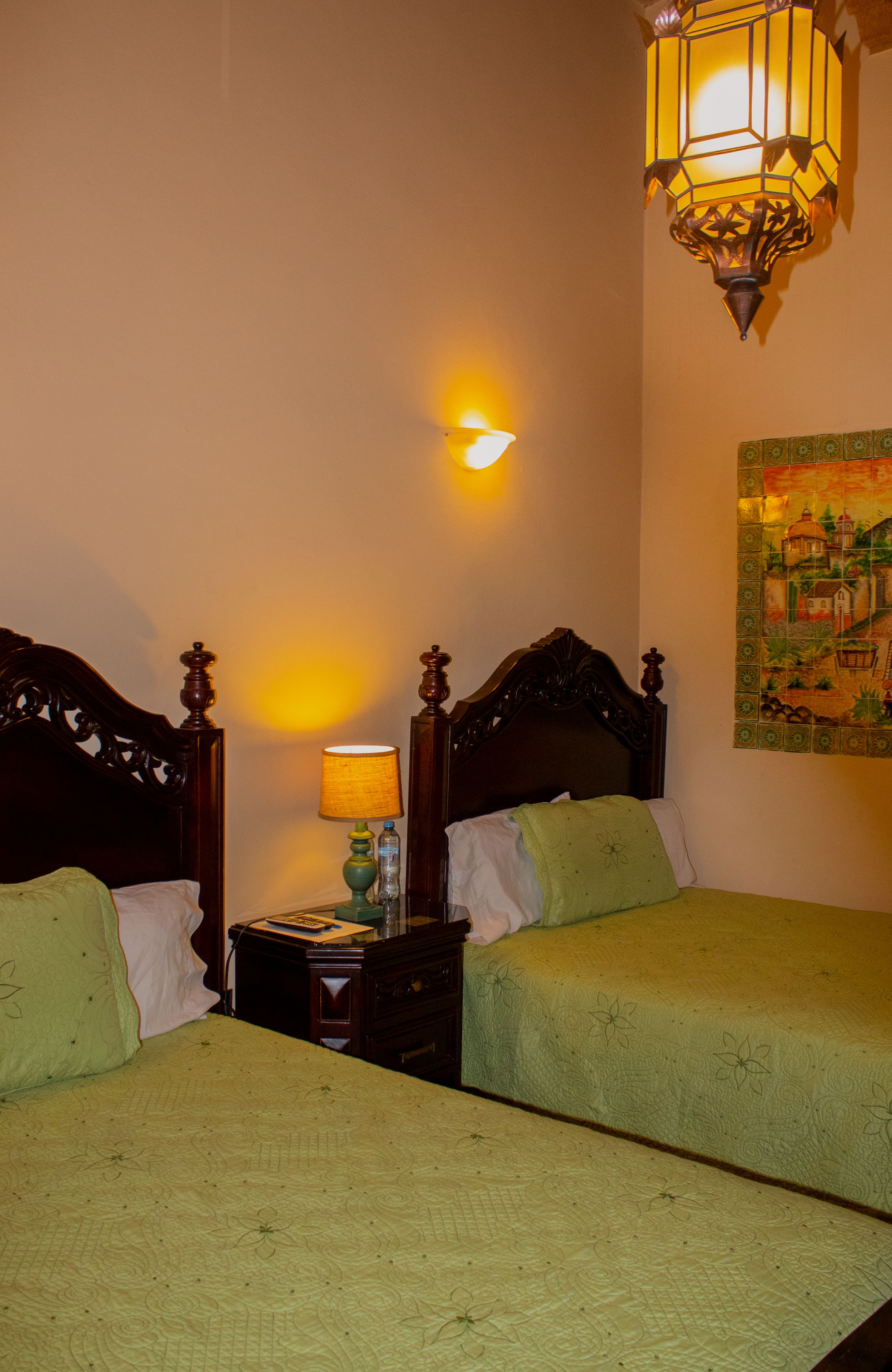 Una habitación con dos camas, una mesita de noche, una lámpara, una luz empotrada en la pared y un mural de azulejos decorativos.