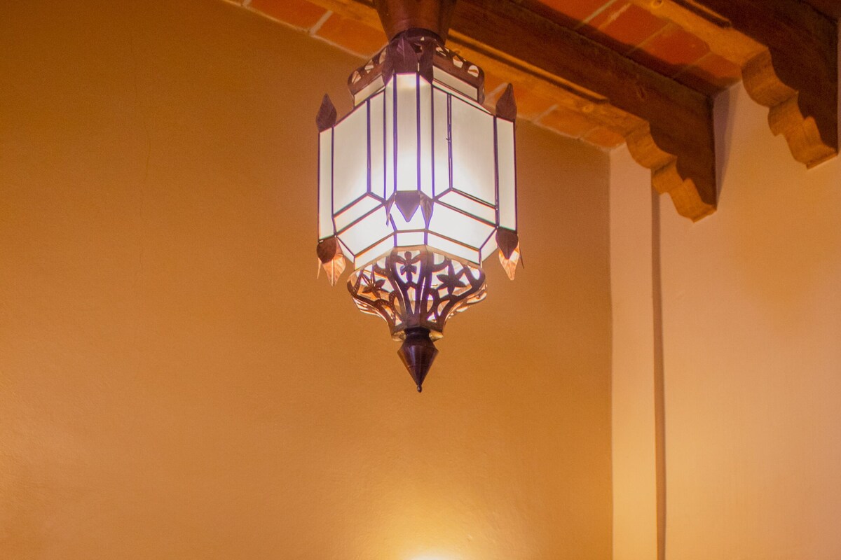 A hotel room with a bed, bedside tables, a lamp, a wall-mounted light, and a decorative wall piece.