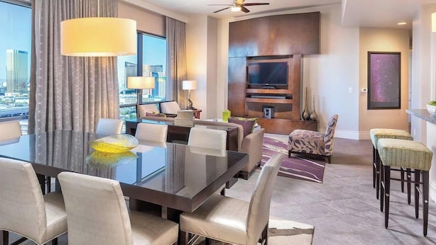 A modern hotel room with a large dining table, comfortable seating, a flat-screen TV, and a city view.